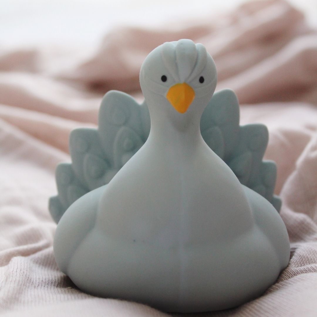 Lyseblå påfugl badedyr i naturgummi fra Natruba, set på lyst sengetæppe,, fås hos Nordic Simply