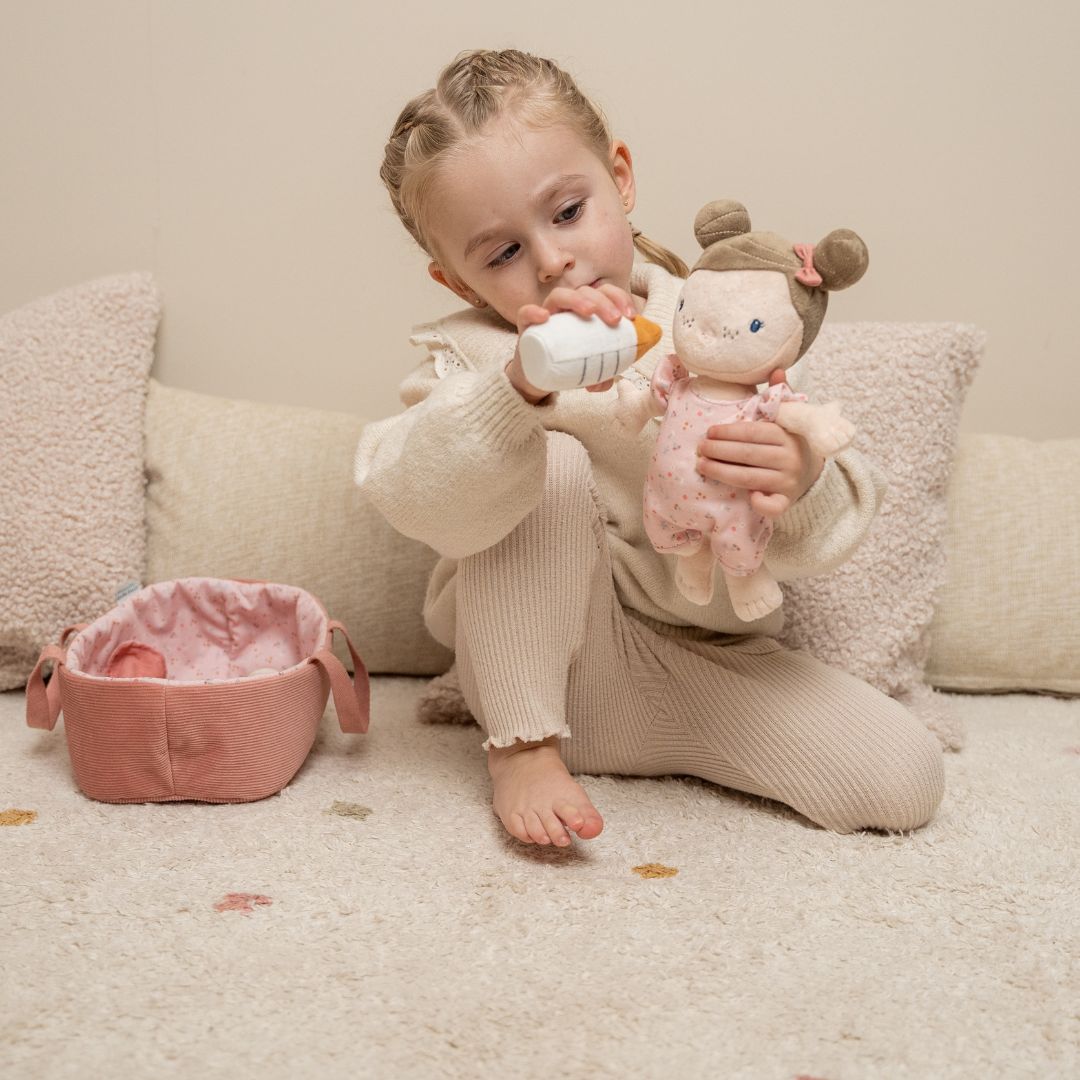 Lille pige leger med blød dukke og sutteflaske i hyggelig stue, perfekt til rolleleg for børn fra 1 år.