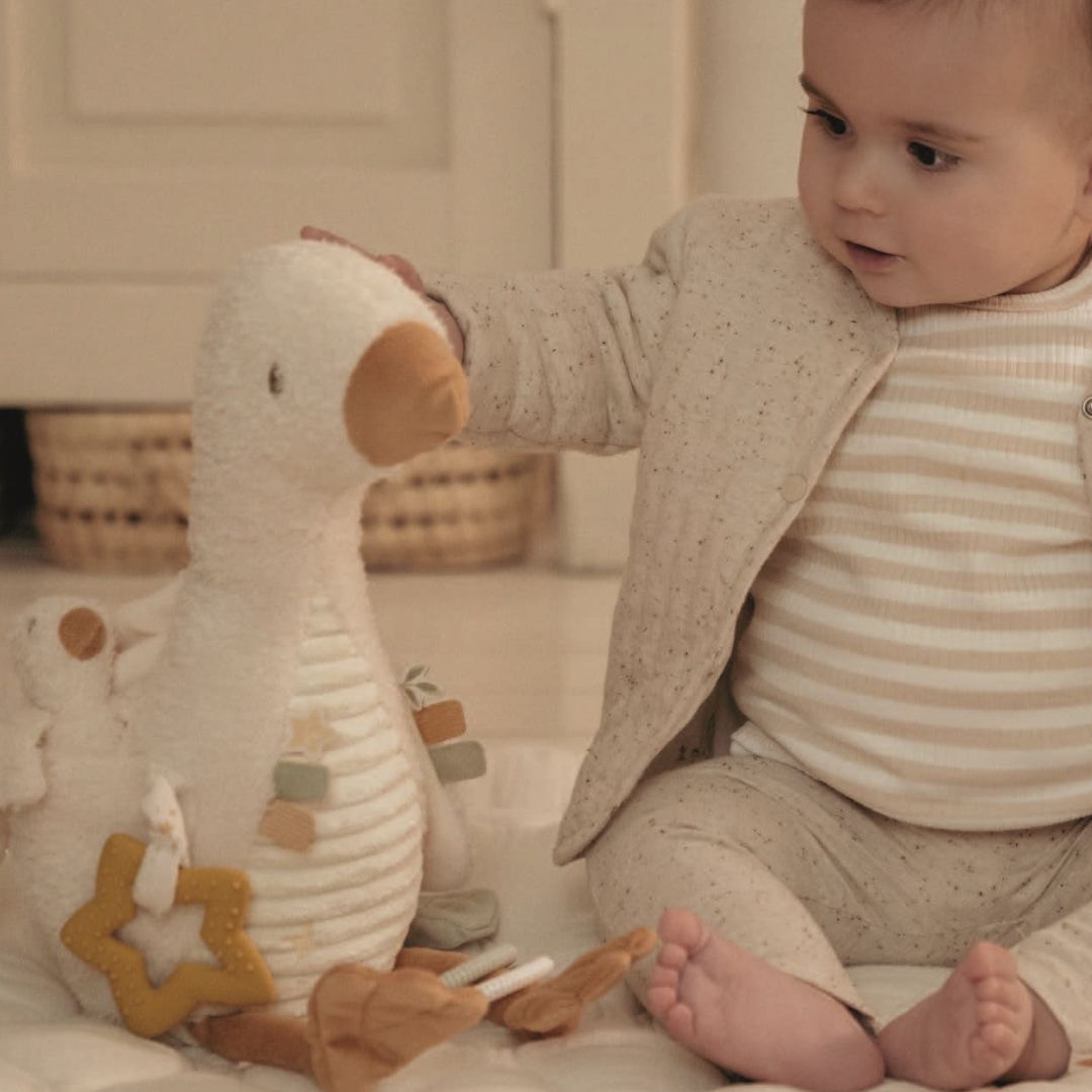 Baby i lyst tøj, sidder på gulv i lyst værelse med hvid gås aktivitetsbamse fra Little Dutch.