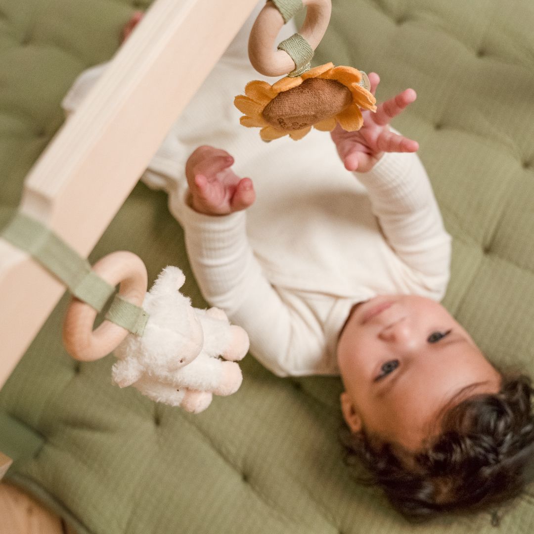 Baby set oppefra ligger under aktivitetsstativ i træ og rører ved en solsikke i plys, der hænger på aktivitetsstativet fås hos Nordic Simply