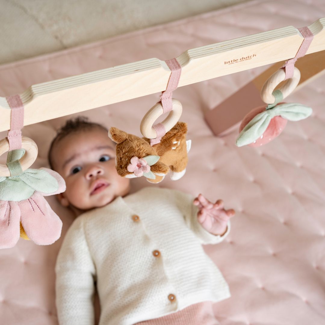 baby pige ligger under aktivitetsstativ i træ med rosa detaljer og kigger på de 3 stykker legetøj, der følger med fra Little Dutch