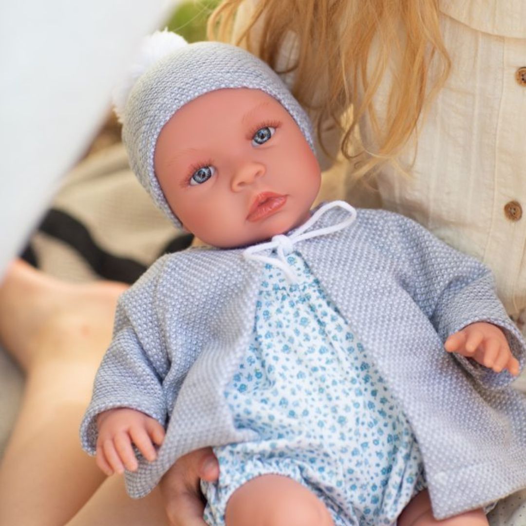 Barn holder stor håndlavet babydukke dreng Leo med klare øjne,i grå jakke og hue samt dragt af lyst liberty stof med små blå blomster, fra Así fås hos Nordic Simply