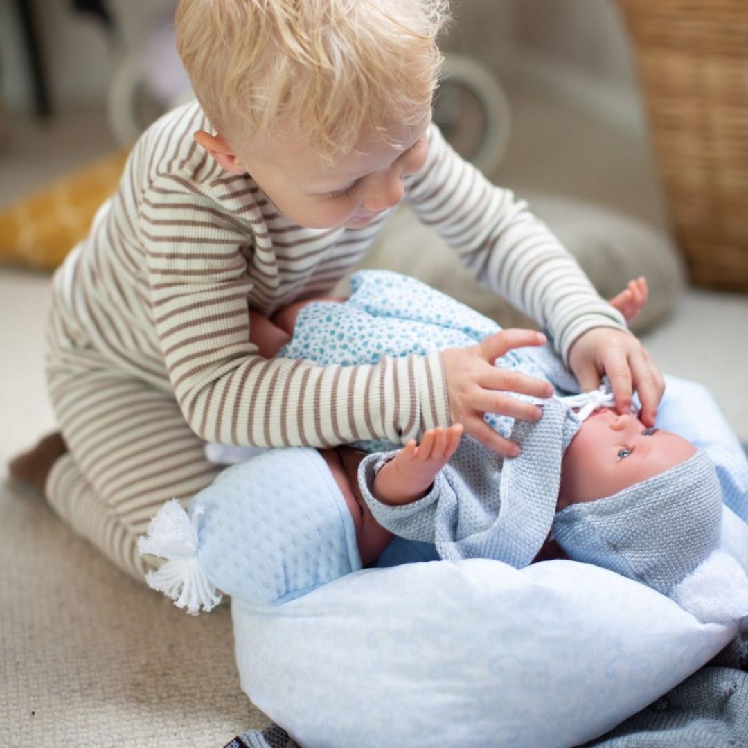 Barn giver babydukke dreng Leo tøj på, grå jakke og hue samt dragt af lyst liberty stof med små blå blomster, fra Así fås hos Nordic Simply