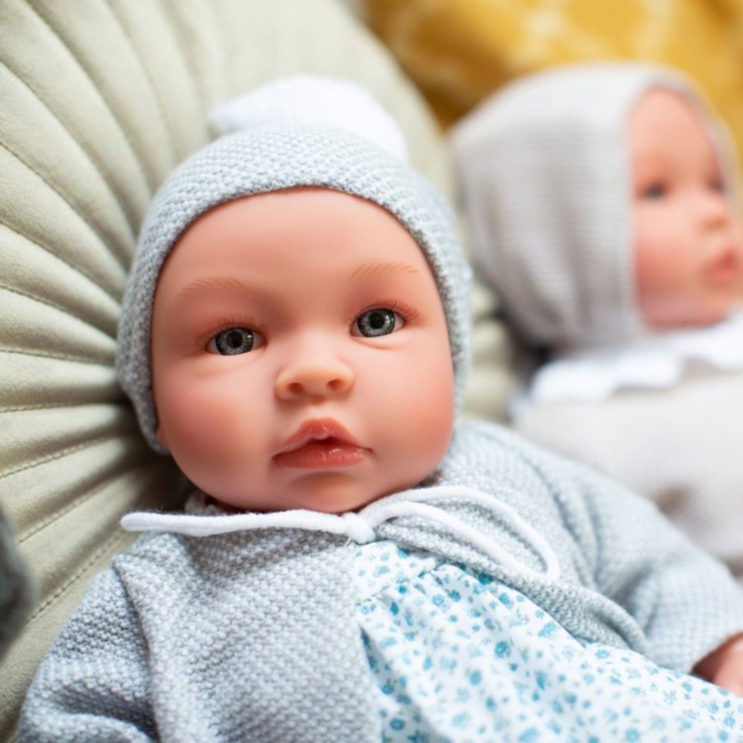 Babydukke dreng Leo med klare øjne ,i grå jakke og hue samt dragt af lyst liberty stof med små blå blomster, kvalitetsdukke fra Así fås hos Nordic Simply