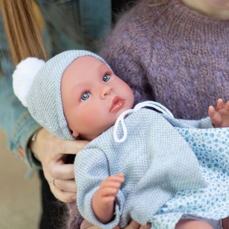 Barn holder stor babydukke Leonora med klare øjne, grå jakke og hue og kjole af lyst liberty stof med små blå blomster, fra Así fås hos Nordic Simply