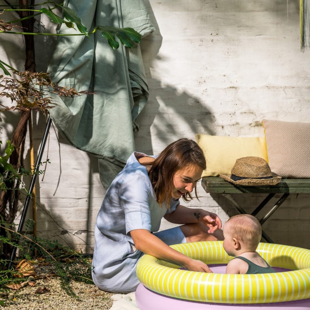 Mor med barn i lille rundt badebassin med to ringe, en rosa og en gul med hvide striber, bassinet har en diameter på 80 centimeter og er derfor til små børn under opsyn.