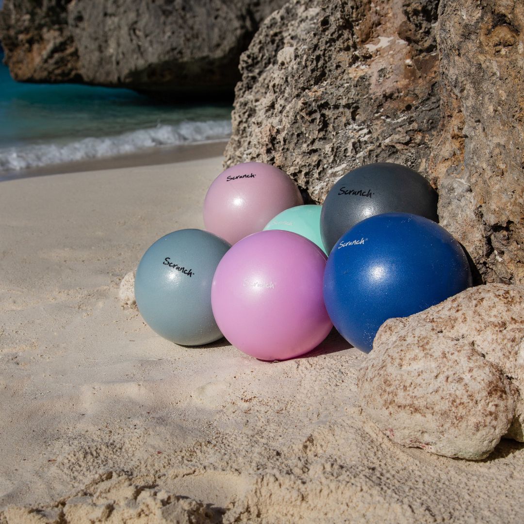 Seks farverige badebolde ved klipper på strand, bagerst ses rosa badebold fra Scrunch, som fås hos Nordic Simply