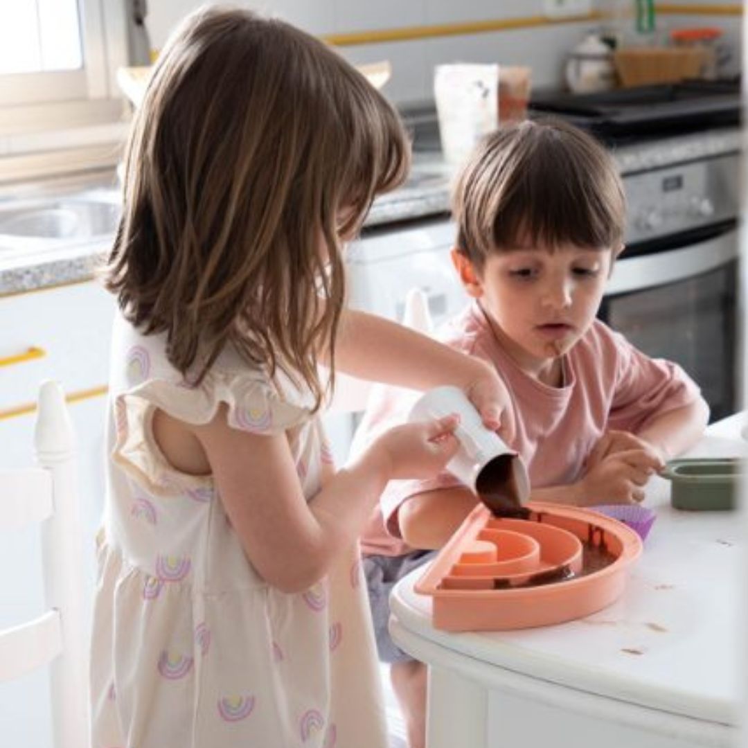 To børn i lyst køkken bager chokoladekage i rosa regnbue bageform fra Dëna fås hos Nordic Simply