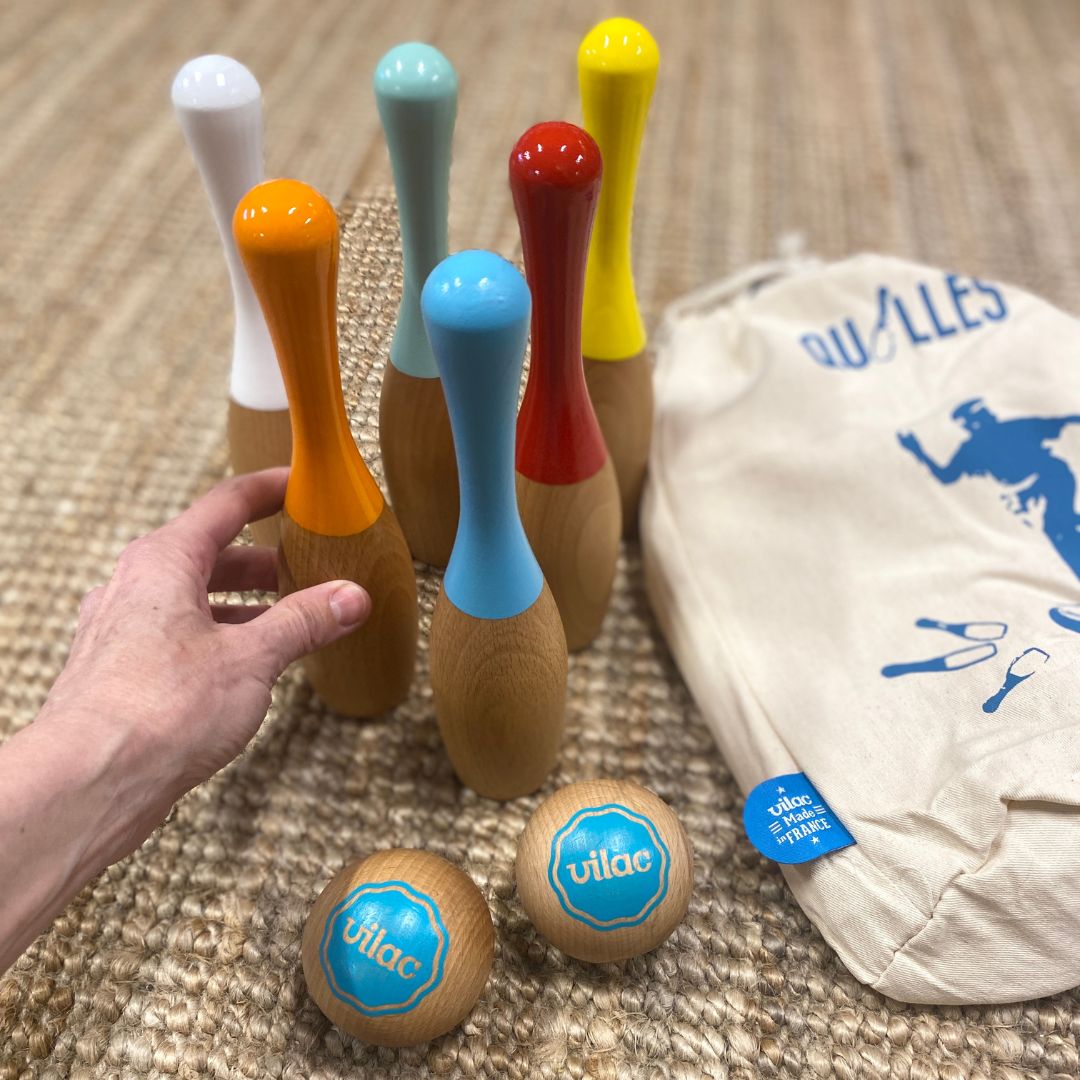 Hånd rører ved kegle fra bowlingsæt i træ i flotte farver og to kugler i træ, ligger på tæppe med stofpose ved siden af.