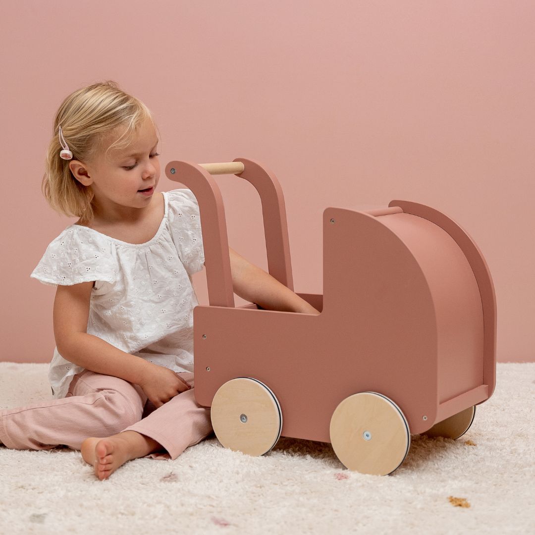 Pige sidder ved rosa dukkevogn i træ fra Little Dutch, i værelse med lyst gulvtæppe og rosa væg, fås hos Nordic Simply