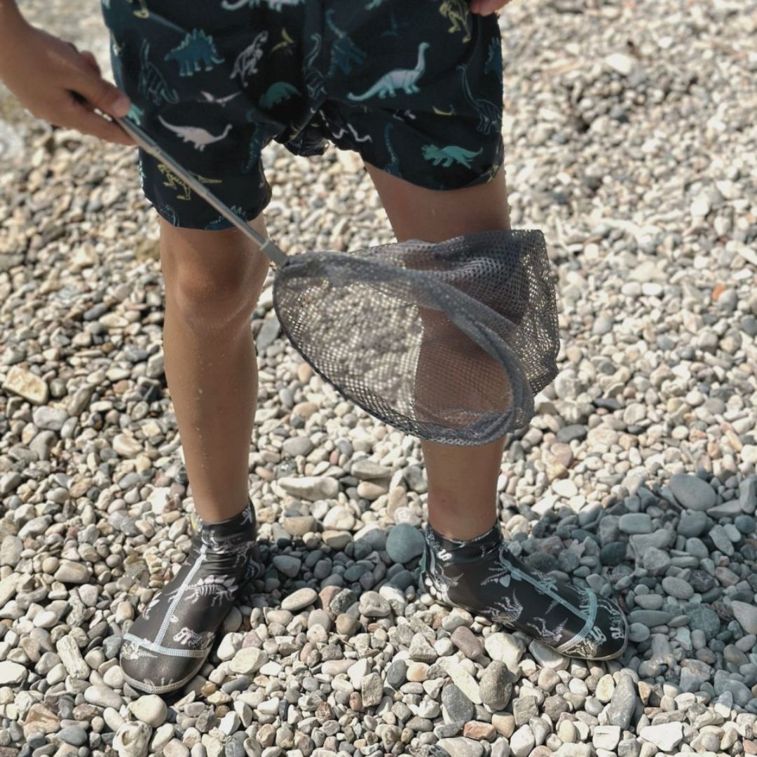 Hånd holder grå fiskenet på teleskopstang på stenstrand, fås hos Nordic Simply