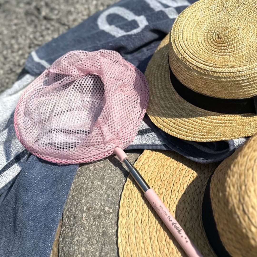 Rosa fiskenet på teleskopstang ligger på strandhåndklæde ved siden af to solhatte.