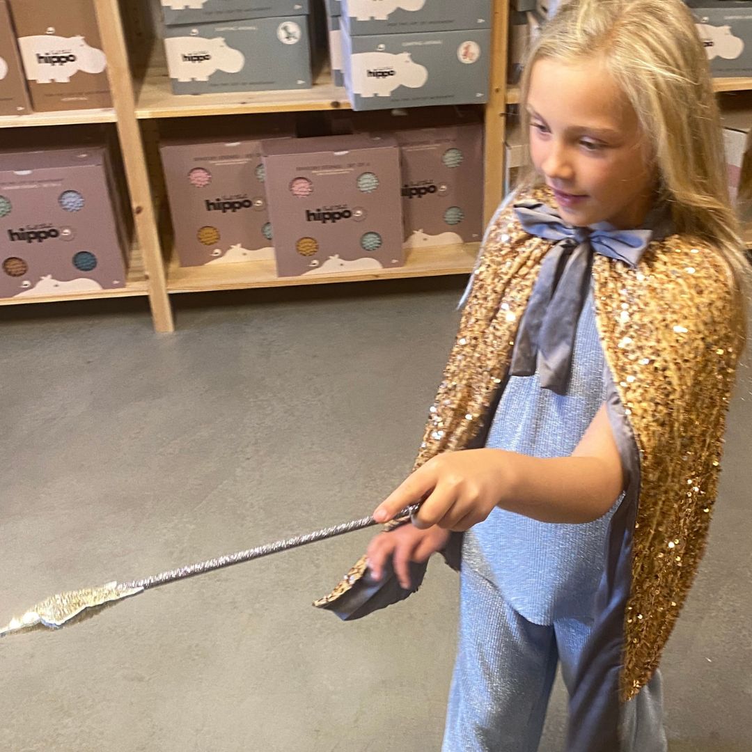 pige i lang guldkappe med pailletter kaster en trylleformular med tryllestav i guld . begge dele fra Mimi & Lula fås hos Nordic Simply