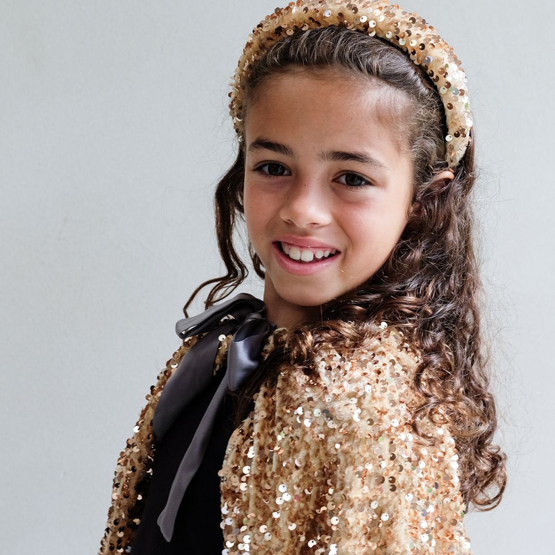 Pige smiler i lang guldkappe og krone, der skinner med tusindvis af pailletter, perfekt udklædning fra 3 til 8 år.