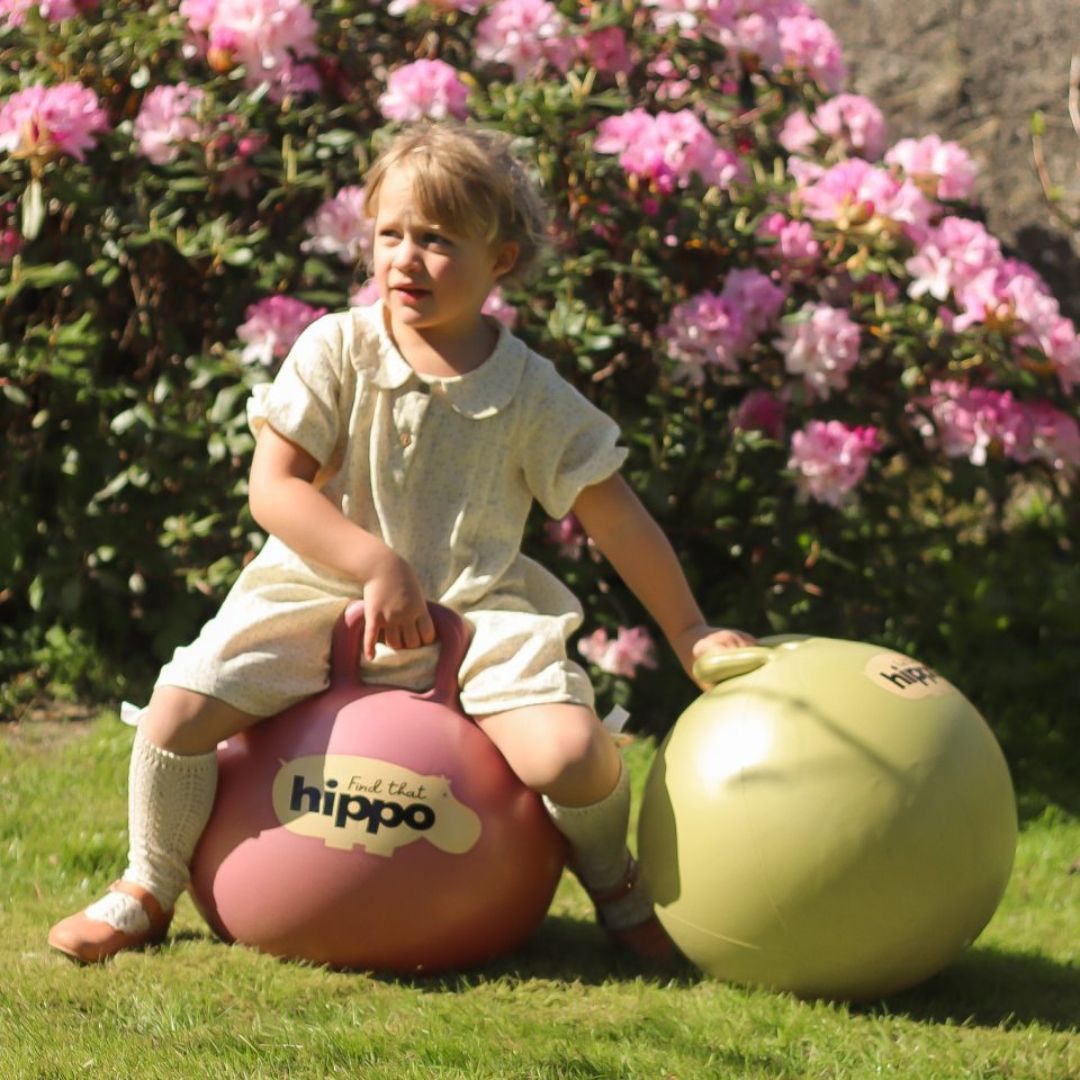 Pige med lange hvid strømper og lys kjole sidder på rosa hoppebold og holder hånd på grøn hoppebold fra Find that Hippo, fås hos Nordic Simply.