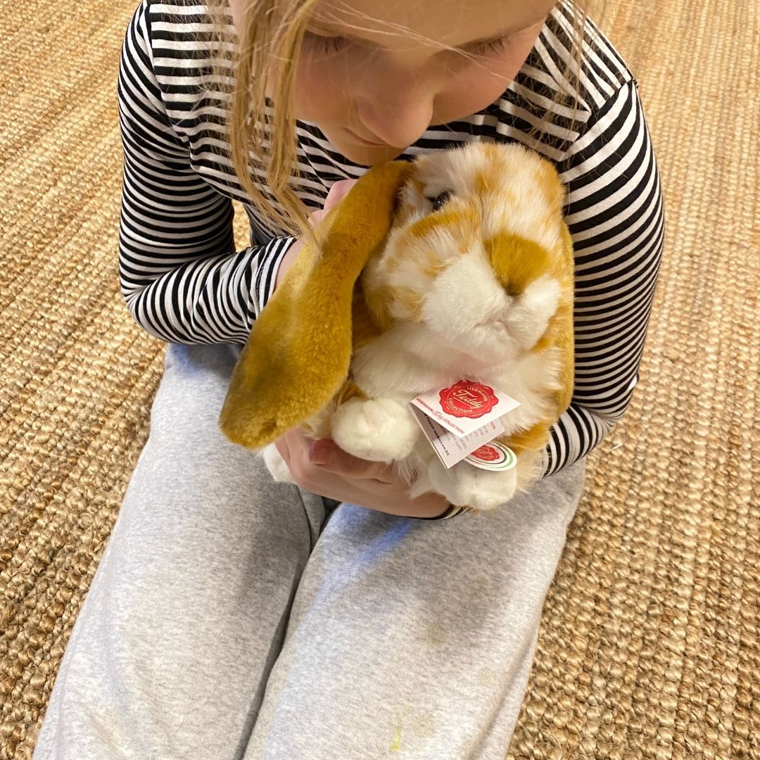 Barn sidder på lysebrunt tæppe og holder brun-hvid vædder kanin bamse, som ægte kæledyr - perfekt til rolle- og fantasileg, fra Teddy Hermann fås hos Nordic Simply