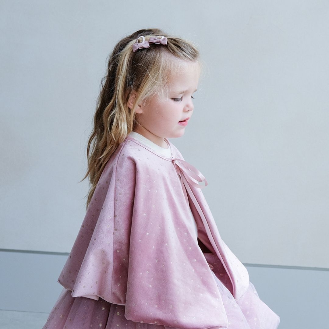 Pige sidder, set fra siden, i rosa kappe i velour dekoreret med små guldstjerne og bindebånd, smuk udklædning i høj kvalitet til børn fra 3 år og op fra Mimi & Lula fås hos Nordic Simply