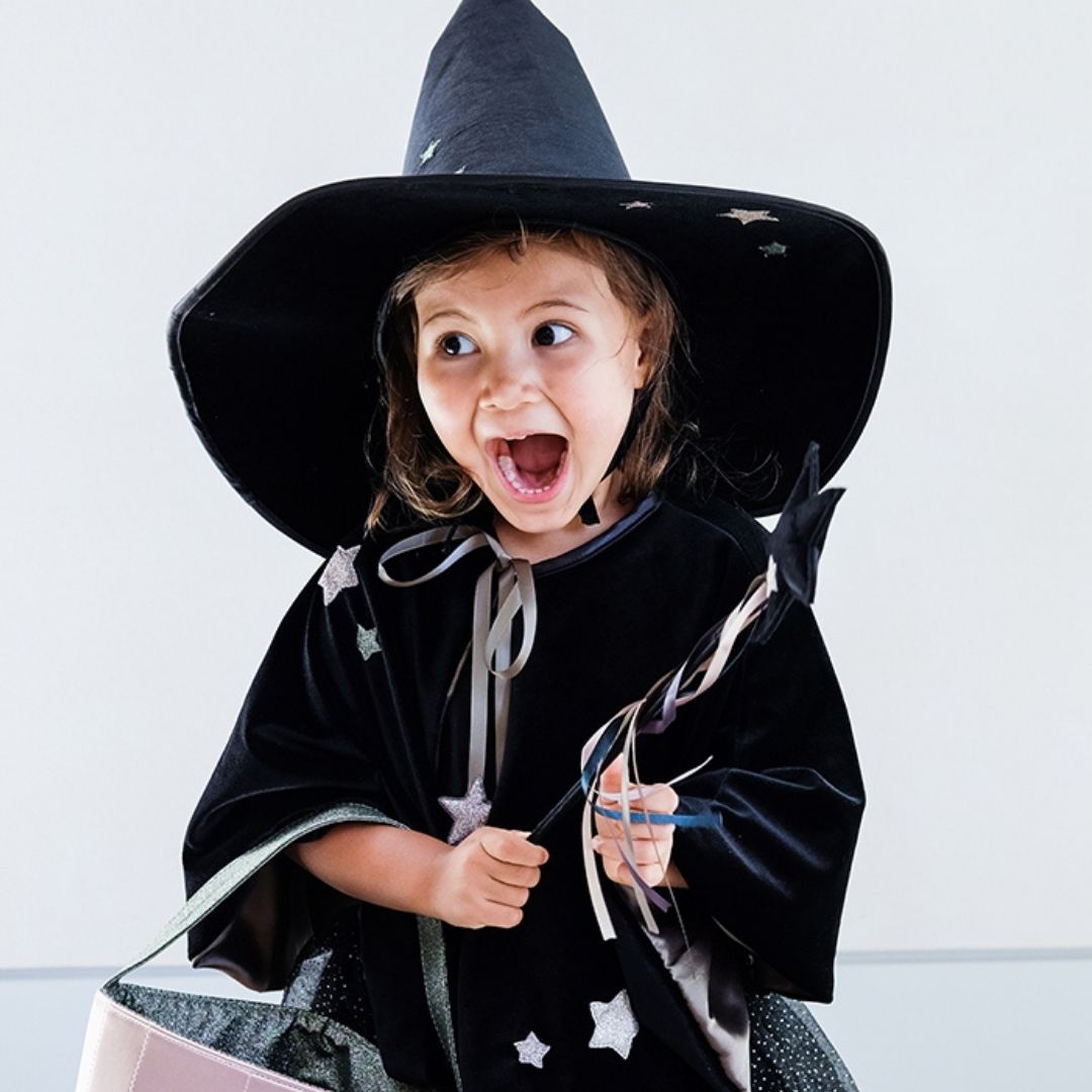 Pige med sort heksehat, tryllestav og kort sort kappe i velour med satinbindebånd, knaplukning og sølvstjerner, flot udklædning til børn fra 3 år og op fra Mimi & Lula fås hos Nordic Simply