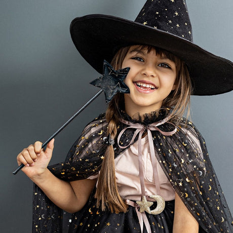 Pige med høj heksehat, tryllestav og lang sort kappe i tyl med rosa satinbindebånd , knaplukning og små guldstjerner og måner, smuk udklædning til børn fra 3 år og op fra Mimi & Lula fås hos Nordic Simply