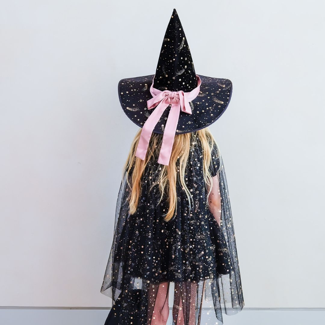 Pige set bagfra med sort heksehat og lang sort kappe i tyl med guldstjerner og rosa bindebånd. Magisk kappe til barn 3-8 år. Vær fe, heks, prinsesse, tryllekunstner eller lige den du vil.