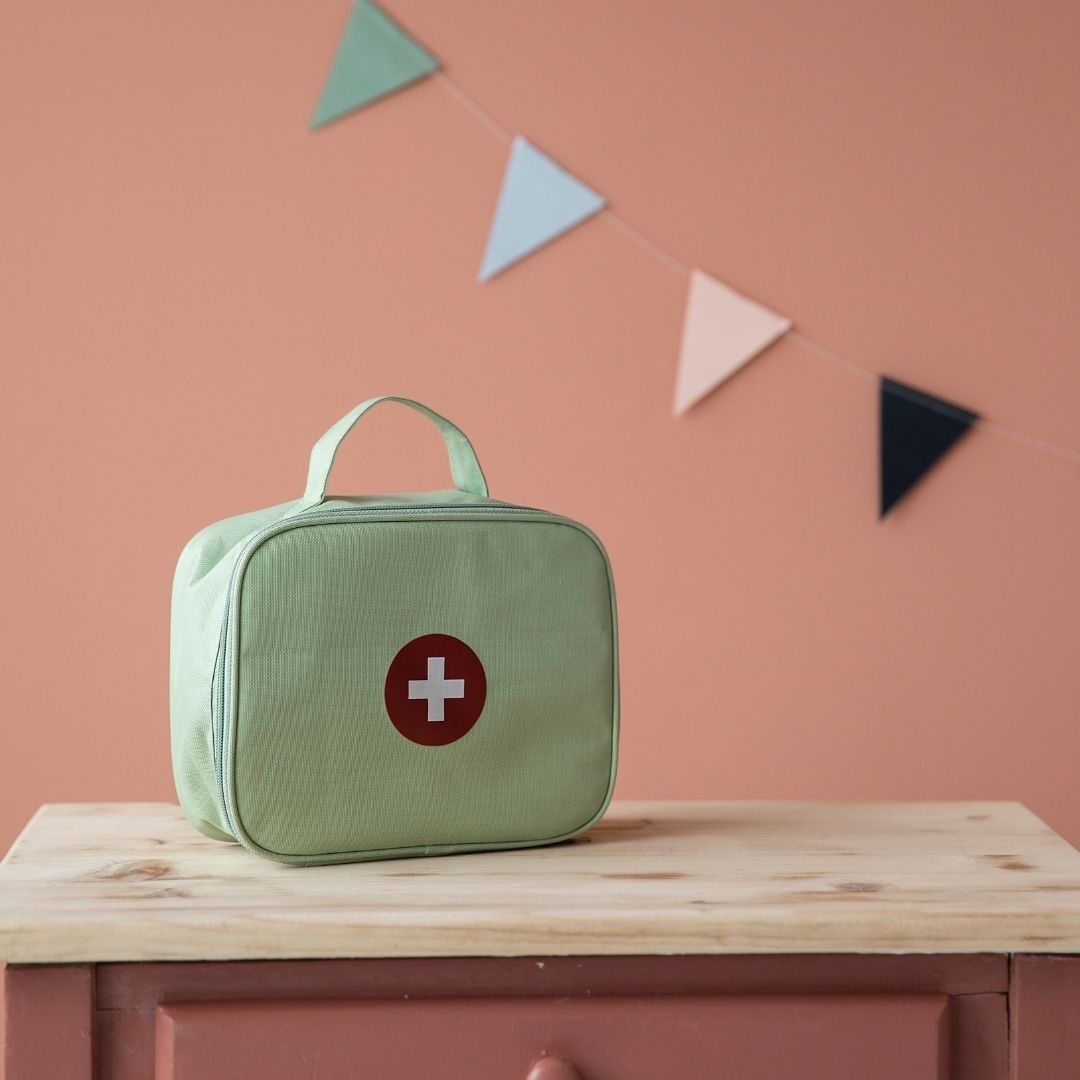 Grøn lægetaske fra Little Dutch med rød cirkel med hvidt kors, står på bord med rosa baggrund.