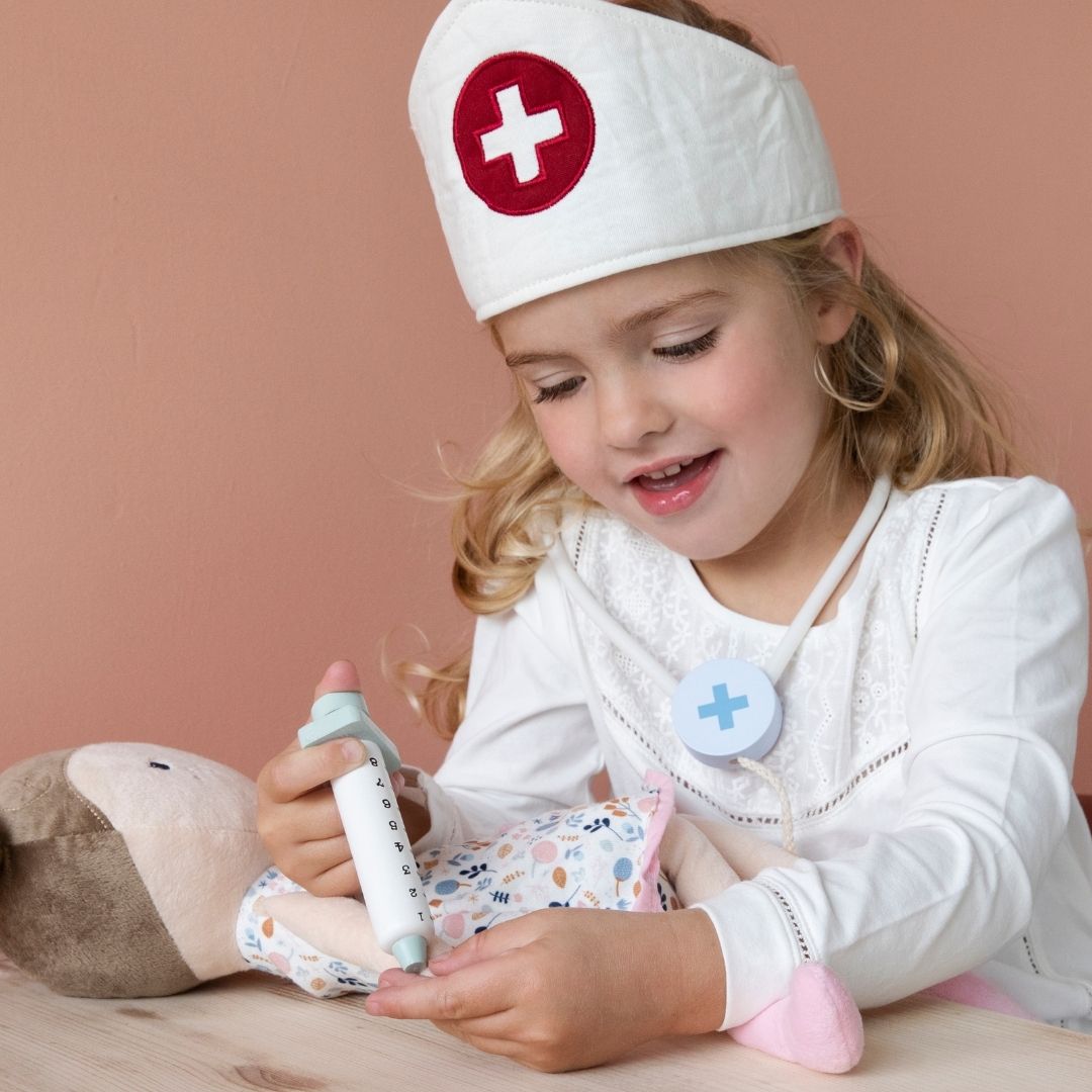 Pige med læge-hat og stetoskop om halsen giver sin dukke en indsprøjtning og leger læge med lægesæt fra Little Dutch på rosa baggrund, fås hos Nordic Simply