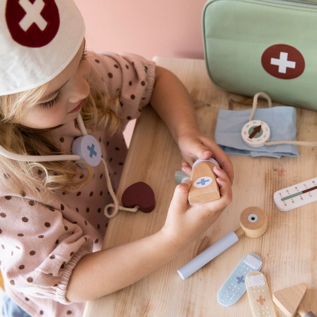 Pige ved bord med stetoskop i træ om halsen, leger doktor med lægesæt i træ fra Little Dutch, her ses forskellige dele som grøn lægetaske, blodtryksmåler, termometer, plaster og salve, legetøjs lægesæt i bedste kvalitet, fås hos Nordic Simply