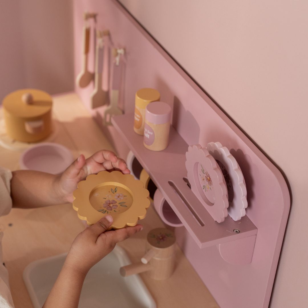 Pink legekøkken med tilbehør set oppefra med små hænder, der holder blomstrede tallerkener