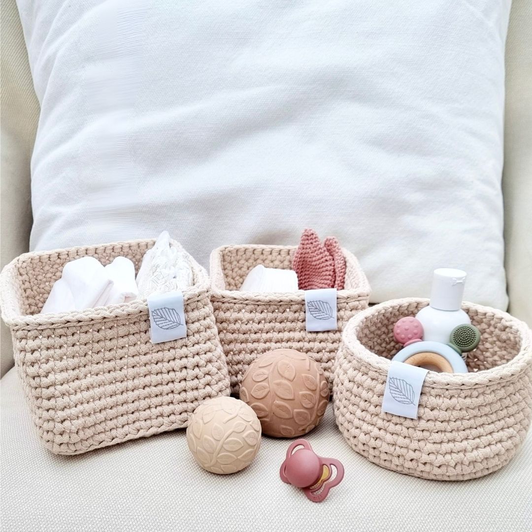 Tre puslekurve i lyse farver med rosa baby-produkter i, står på pude. Puslekurvesættet er håndlavet i Danmark, fås hos Nordic Simply