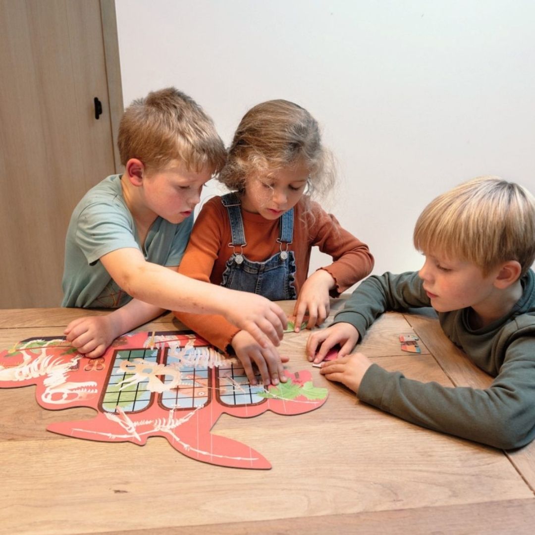 Tre børn ved bord lægger puslespil med dinosaurer, der kan vendes, her lægges bagsiden, 100 brikker fra Scratch fås hos Nordic Simply