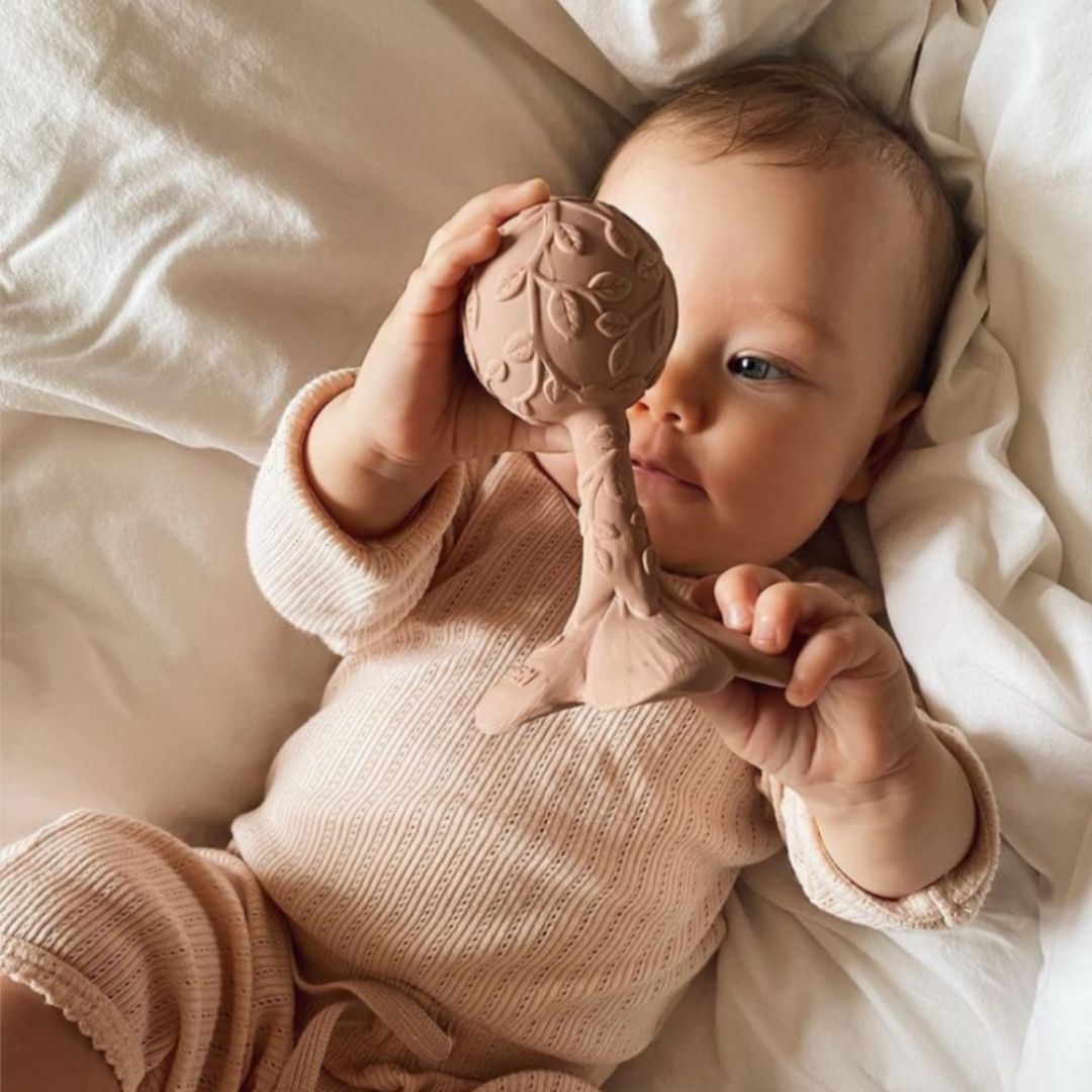 Baby undersøger rosa rangle og bidering i et, formet som en plumeria, med teksturer, fra Natruba, fås hos Nordic Simply