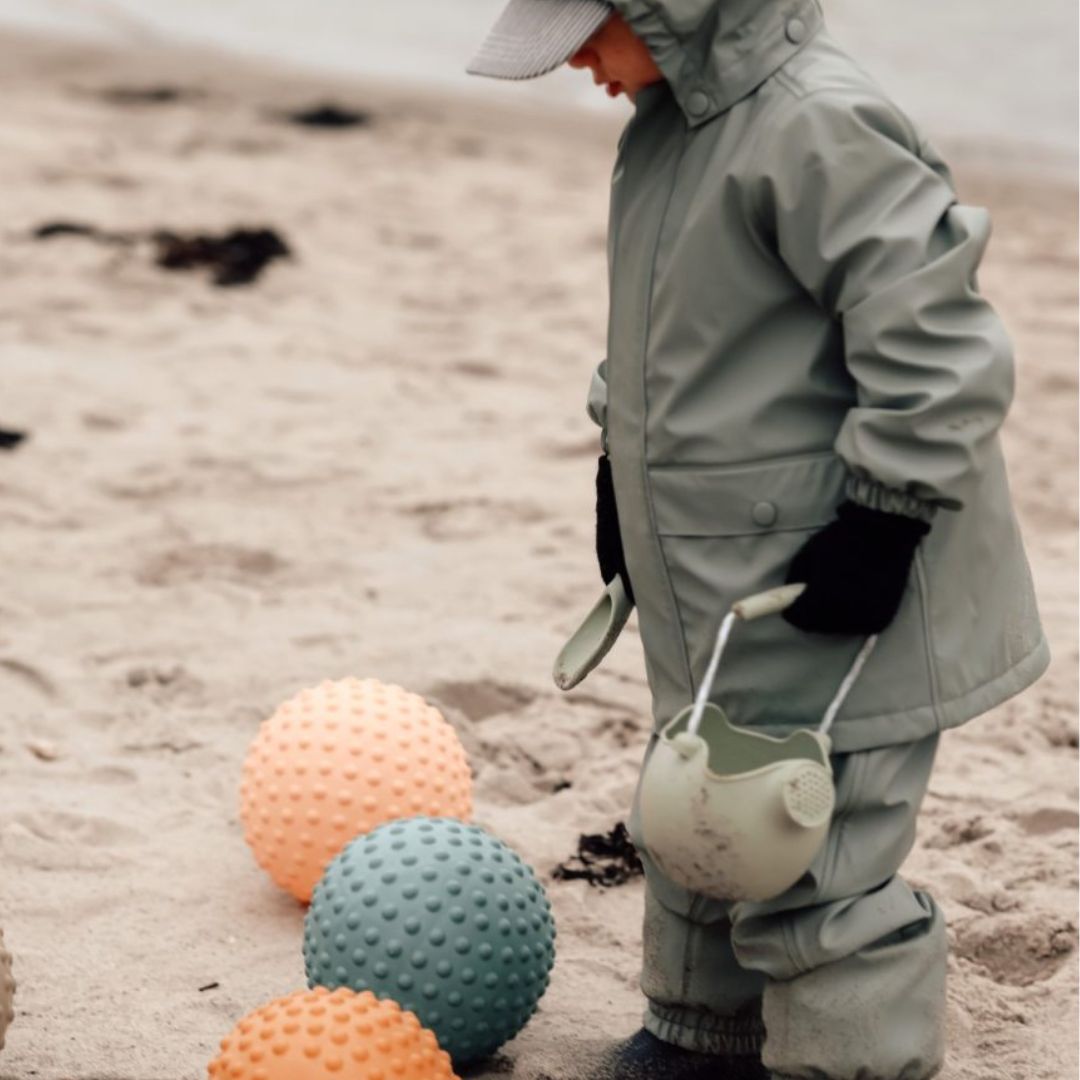 Barn i regntøj står på stranden ved sansebolde i farverne orange og blå.