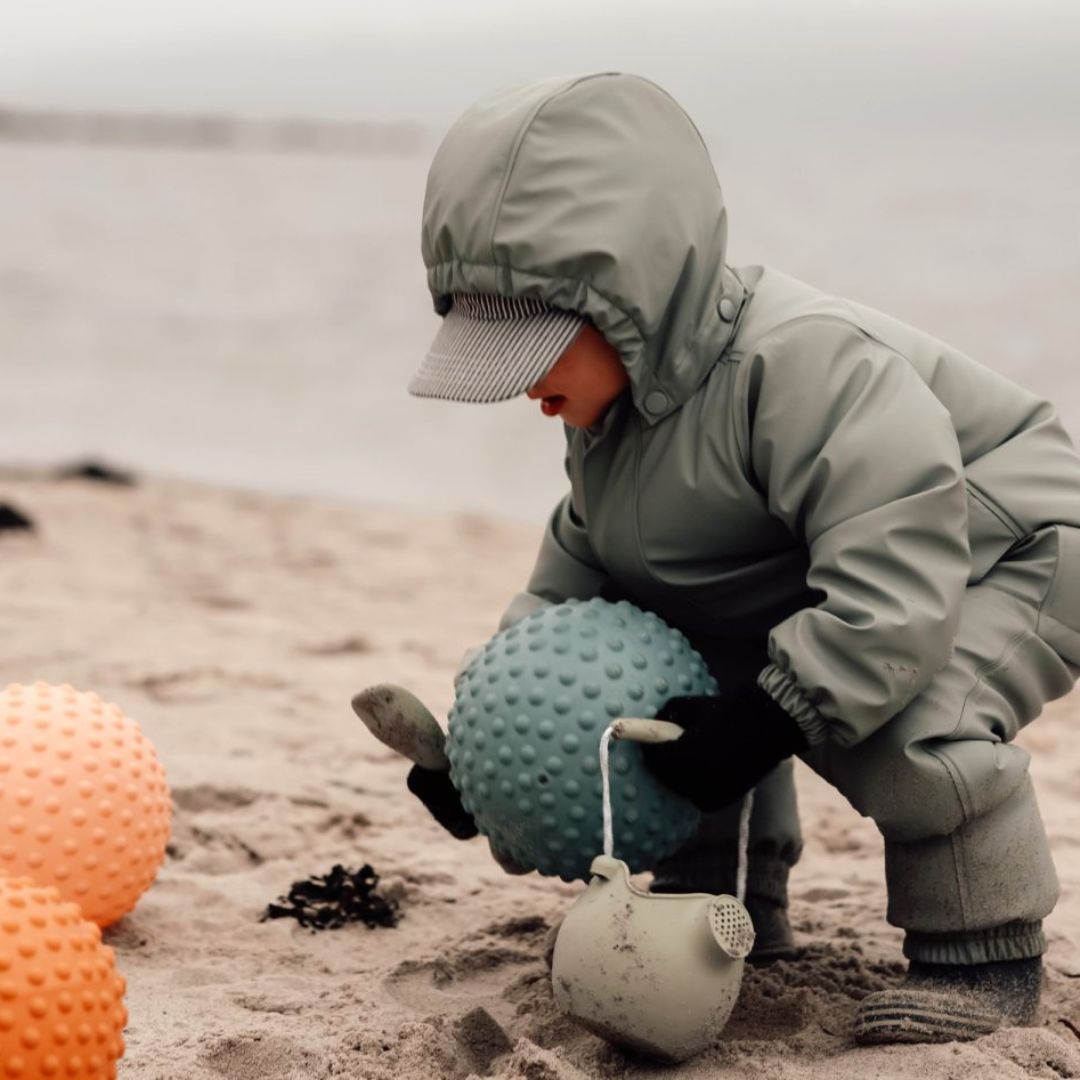 Barn i gråt regntøjj leger med en stor blå sansebold på stranden, omgivet af flere farverige sansebolde.