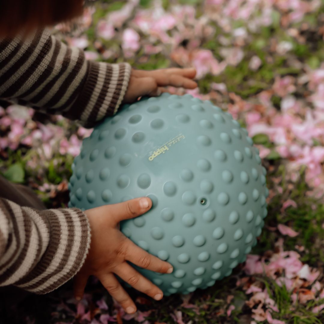Barn griber stor blå sansebold med knopper i støvet blå farve blandt blomsterblade.
