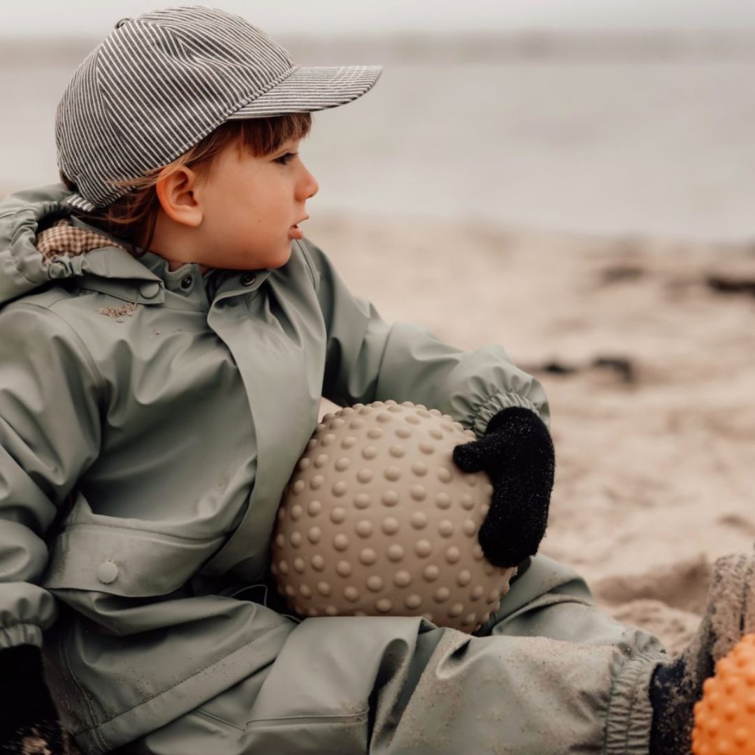 Barn i grøn regntøj holder en grøn sansebold på stranden, perfekt til motorik og sansestimulation.