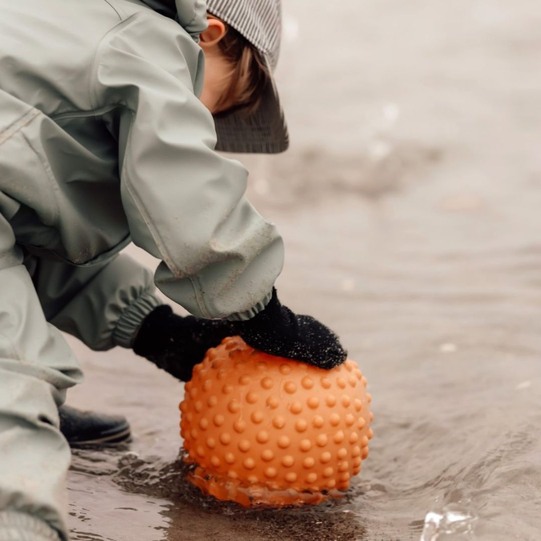 Barn i regntøj, leger på strand med orange sansebold, ideel til sansestimulation og motorisk udvikling.