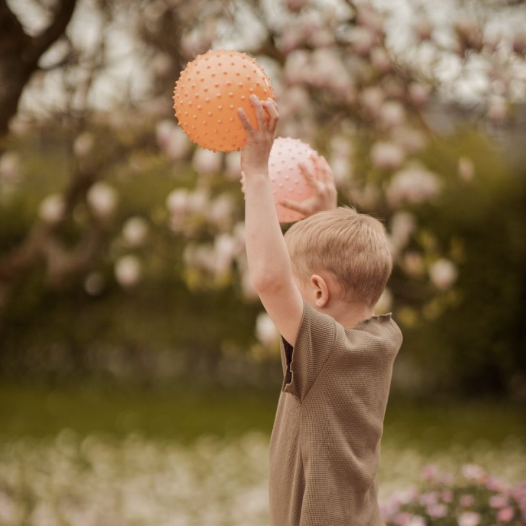 Barn i naturen holder stor orange sansebold, i venstre hånd og stor rosa sansebold i højre hånd. Ideel til sansestimulation og motorisk udvikling til baby og store børn.