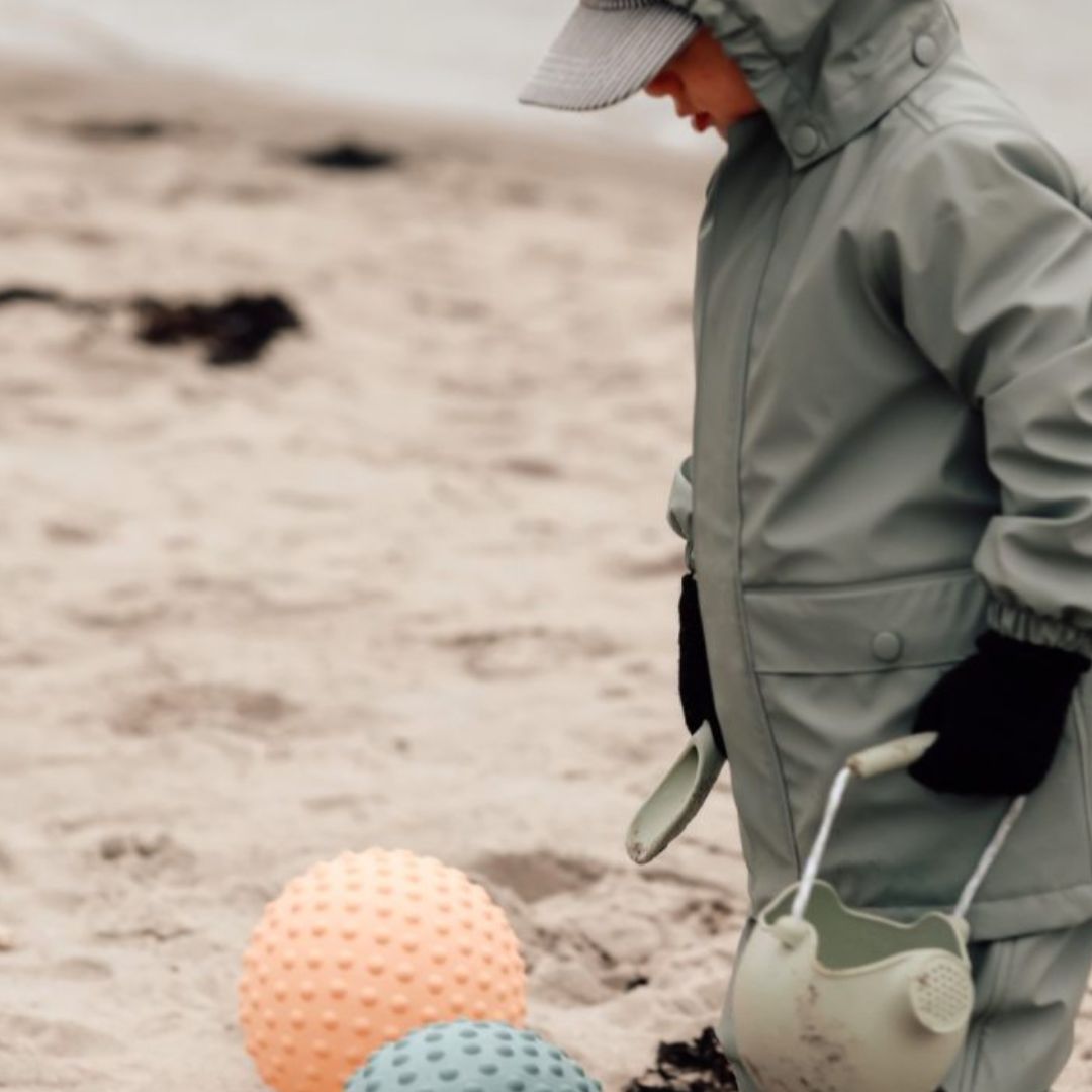 Barn i grå regndragt leger på stranden med stor orange sansebold og grøn sansebold til sansestimulering.