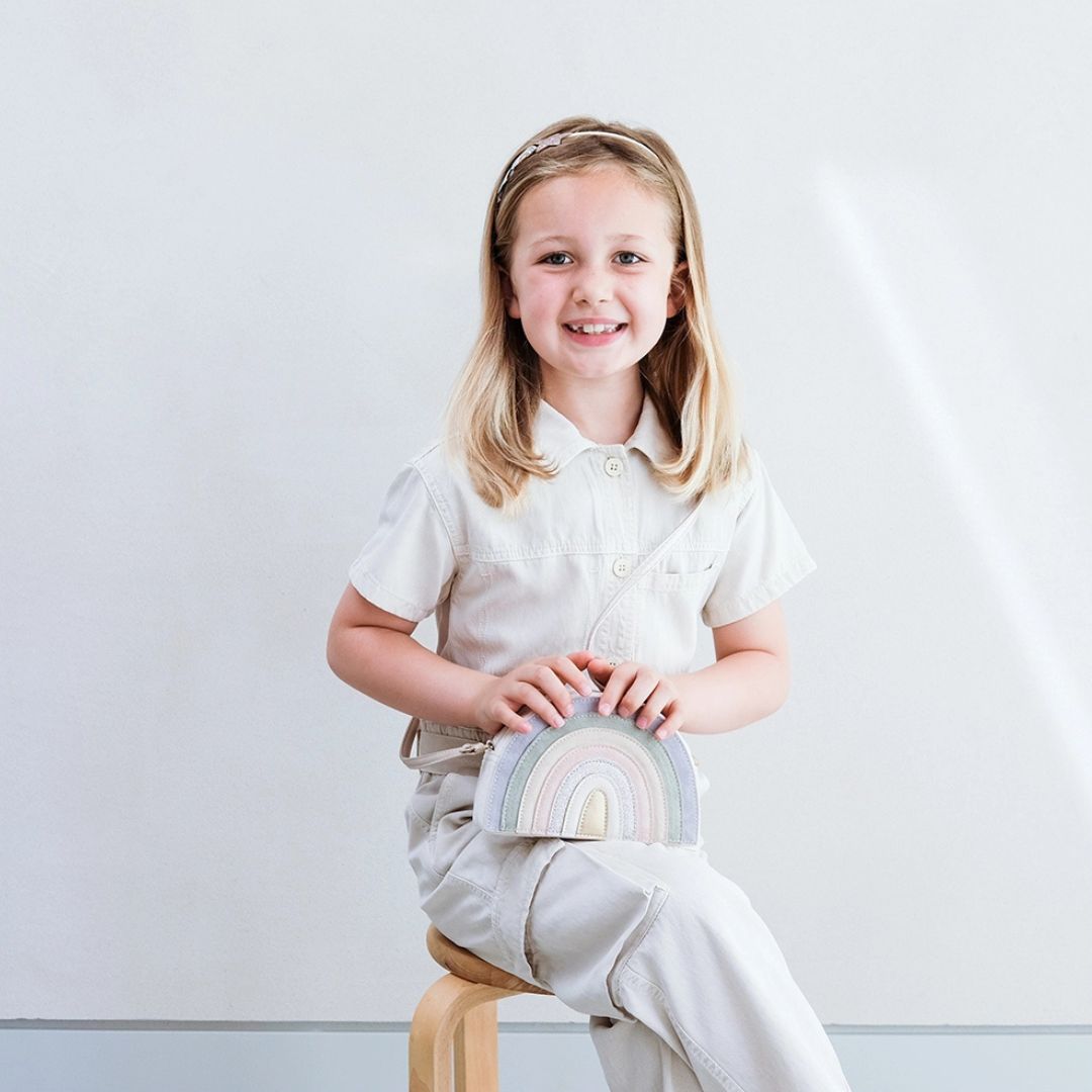 Pige sidder med taske formet som regnbue i lyse pastelsfarver, skuldertaske til børn fra 3 år og op, med aftagelig strop, fra Mimi & Lula fås hos Nordic Simply