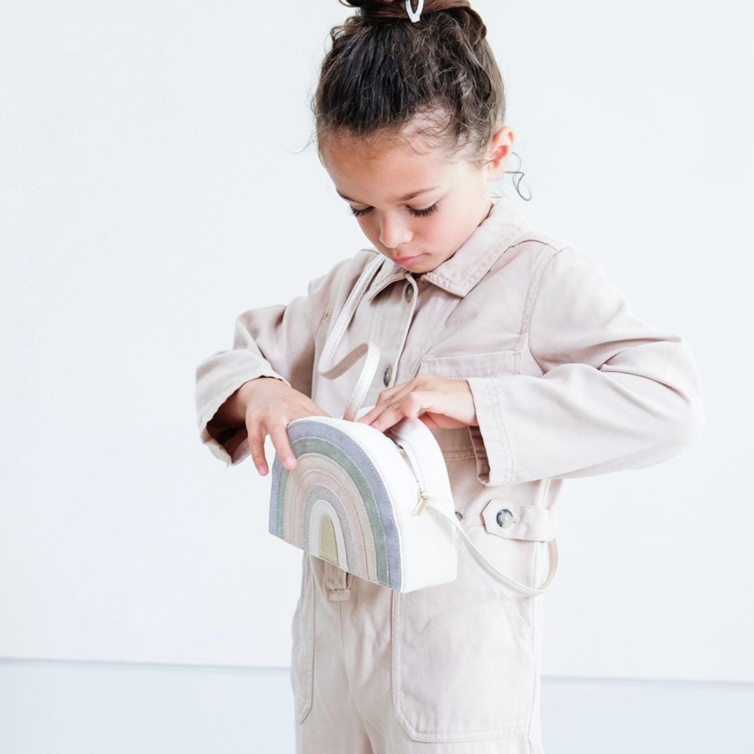 Pigge ser ned i taske formet som regnbue i lyse pastelsfarver, skuldertaske til børn fra 3 år og op, med aftagelig strop, fra Mimi & Lula fås hos Nordic Simply