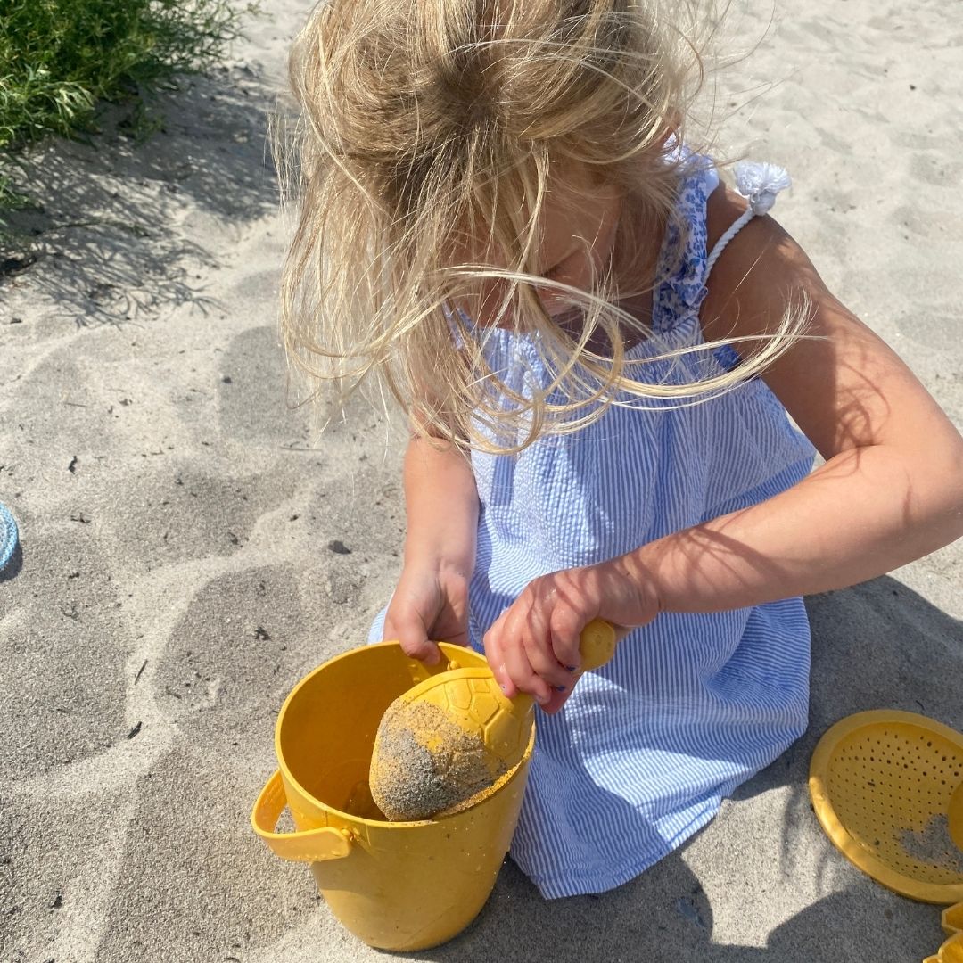 Pige leger i sand med gul spand med si, skovl og to forme, strandsæt i holdbart bioplast fra Lässig, fås hos Nordic Simply