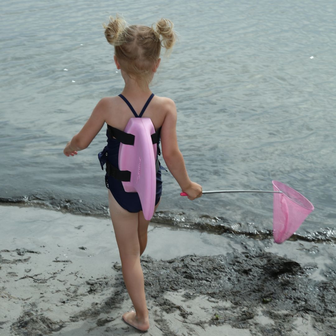 Pige på strand med ryggen til med rosa fiskenet i hånden og SwimFin hajfinne på ryggen. Original SwimFin fås hos Nordic Simply