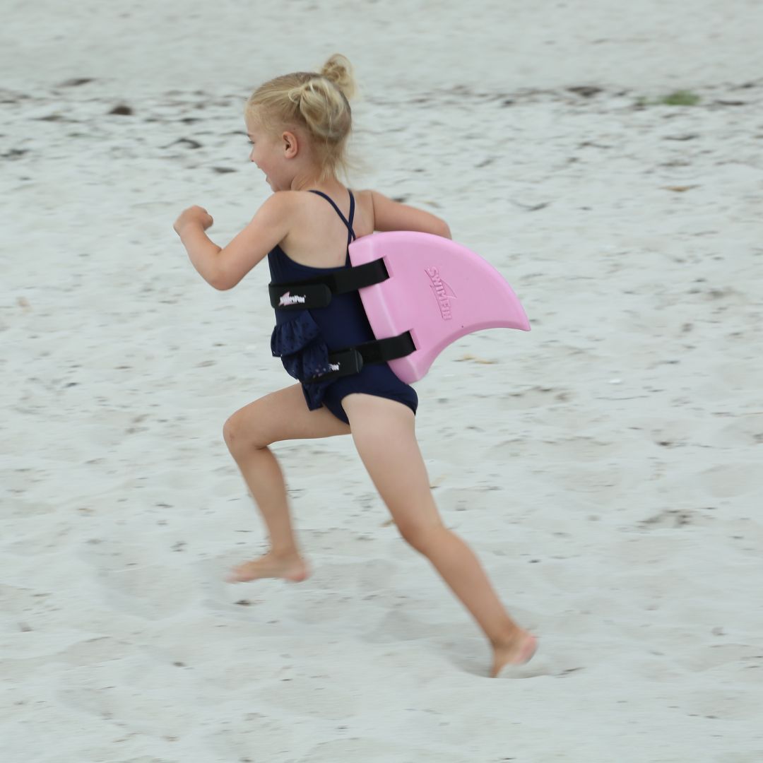 Pige løber på strand med rosa SwimFin hajfinne på ryggen. Original SwimFin fås hos Nordic Simply