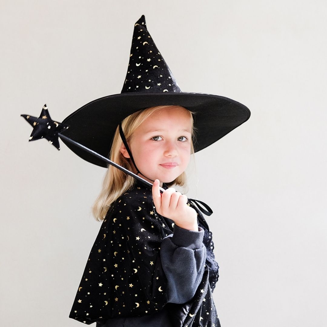 Pige med sort heksehat, kort sort kappe og sort tryllestav med stjerne på toppen i velour med små sølvstjerner, kommer på træpind beklædt med sort satin, perfekt som tilbehør til udklædning som fe, prinsesse, heks, sommerfugl eller andet fra Mimi & Lula fås hos Nordic Simply