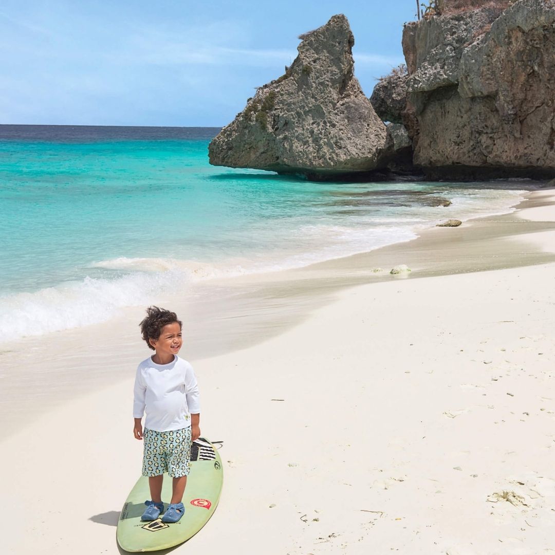 Dreng står på surfboard på hvid samed hvid sandstrand med uv-bluse med lange ærmer str 24 måneder fra Lässig fås hos Nordic Simply