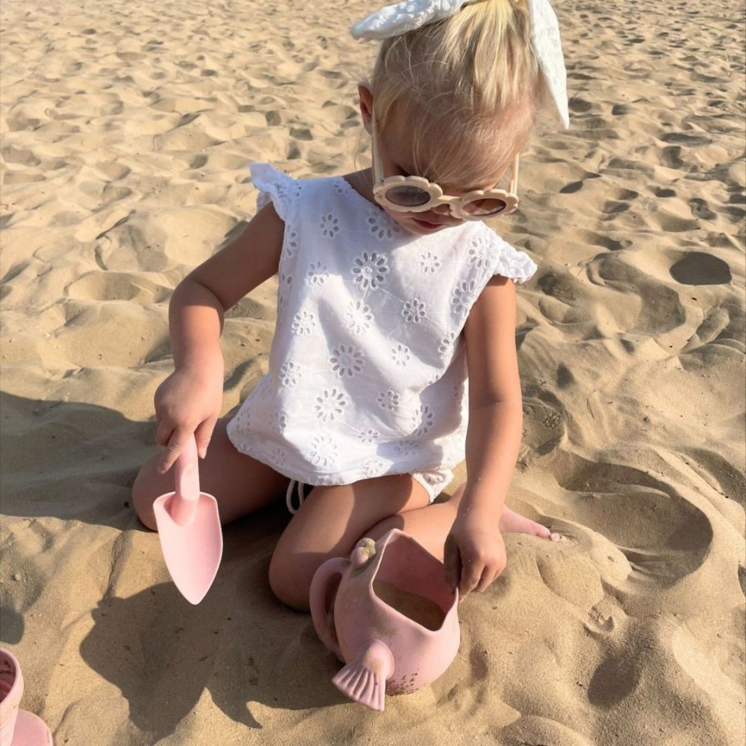 Pige på strand hælder sand i rosa vandkande og skovl i én, formet som en fisk med stor mund, fra Scrunch fås hos Nordic Simply