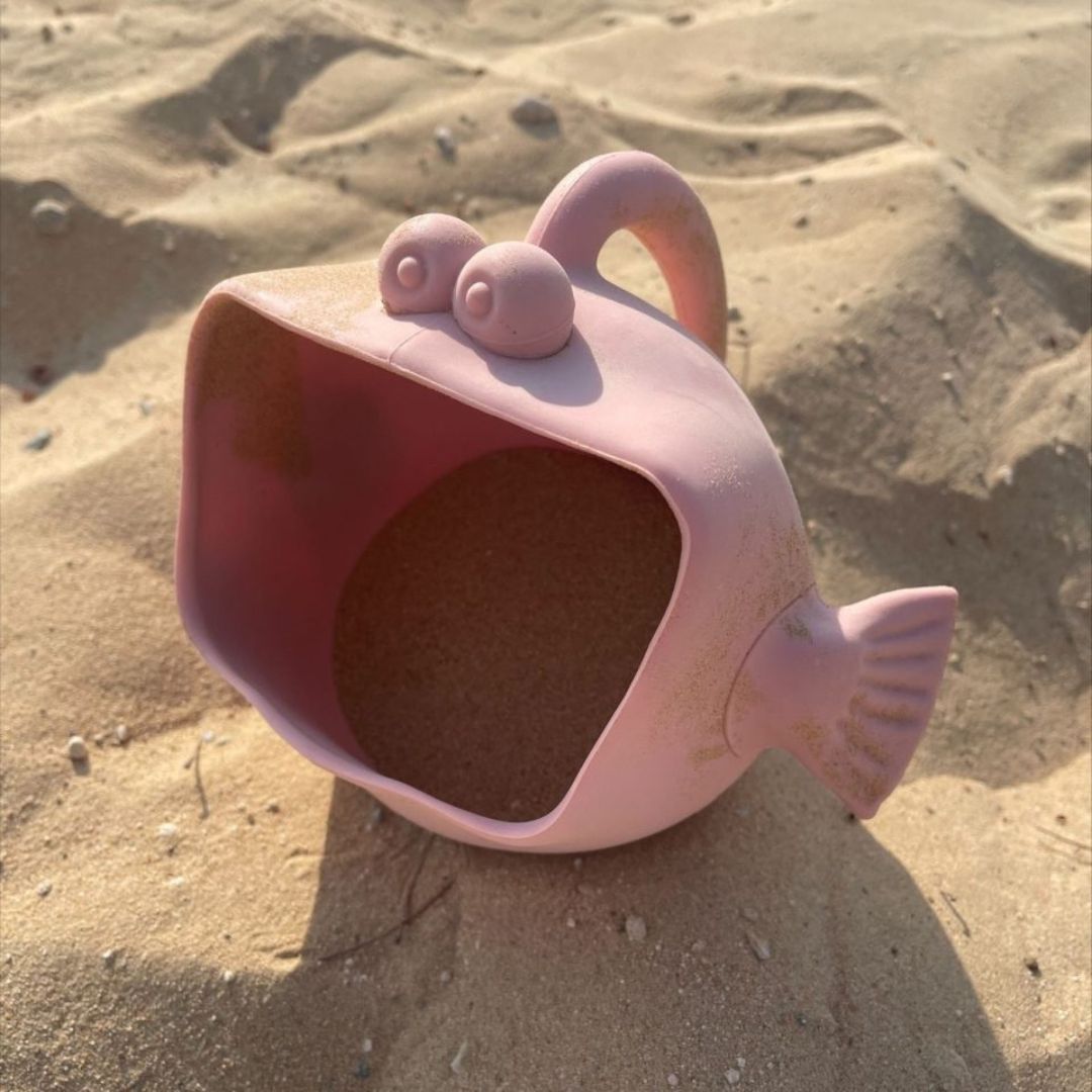 Rosa vandkande og skovl i én, formet som en fisk med stor mund, brug den til vand, sand eller sne, her set i sand, fra Scrunch fås hos Nordic Simply