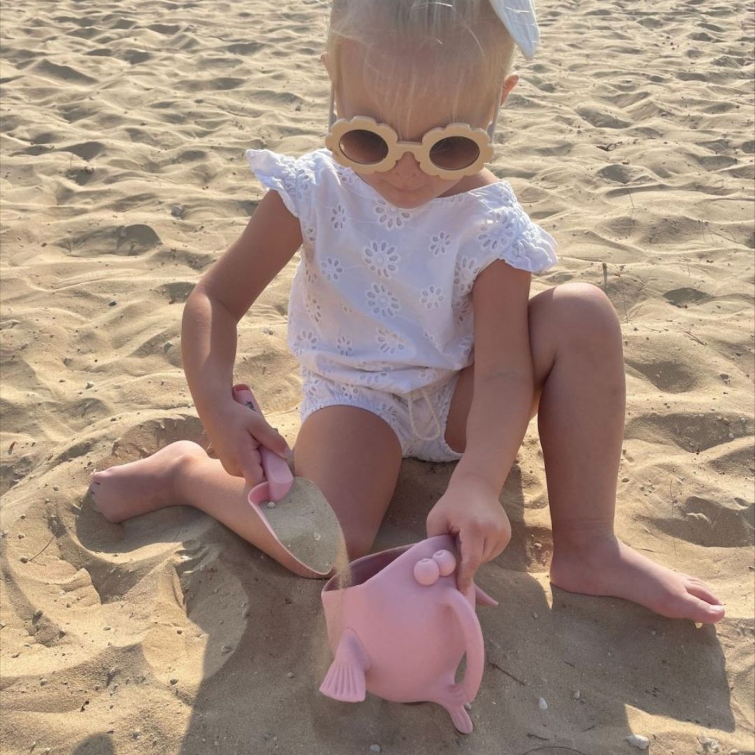 Pige på strand hælder sand i rosa vandkande og skovl i én, formet som en fisk med stor mund, fra Scrunch fås hos Nordic Simply
