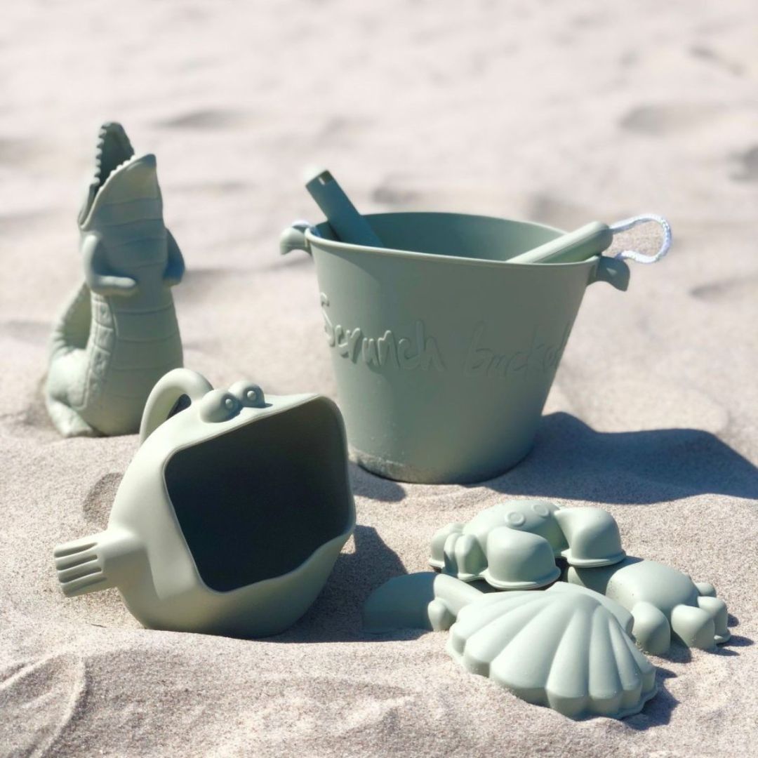 Grøn vandkande formet som en krokodille, set i sand på strand sammen med mere strandlegetøj fra Scrunch, fås hos Nordic Simply