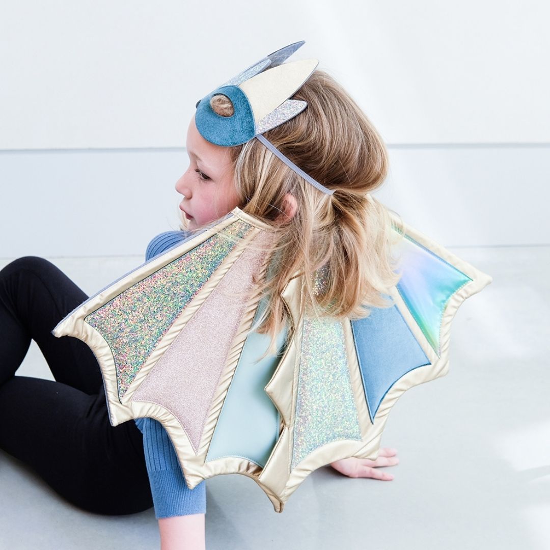 Pige sidder, set bagfra, med dragemaske i panden og vinger på ryggen i pastelfarver med glitter og guld fra Mimi & Lula fås hos Nordic Simply